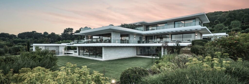 Contemporary luxury villa with white geometric volumes and expansive floor-to-ceiling glass walls at sunset.