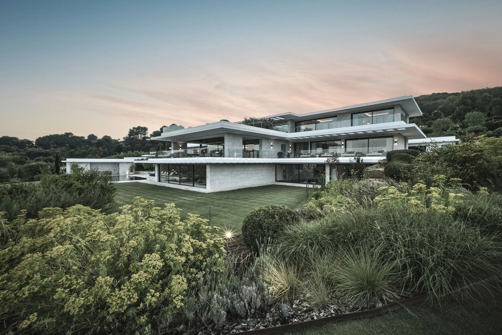 Wide shot of the modern Mediterranean facade with stacked horizontal planes, glass railings, and native shrubbery in the foreground.