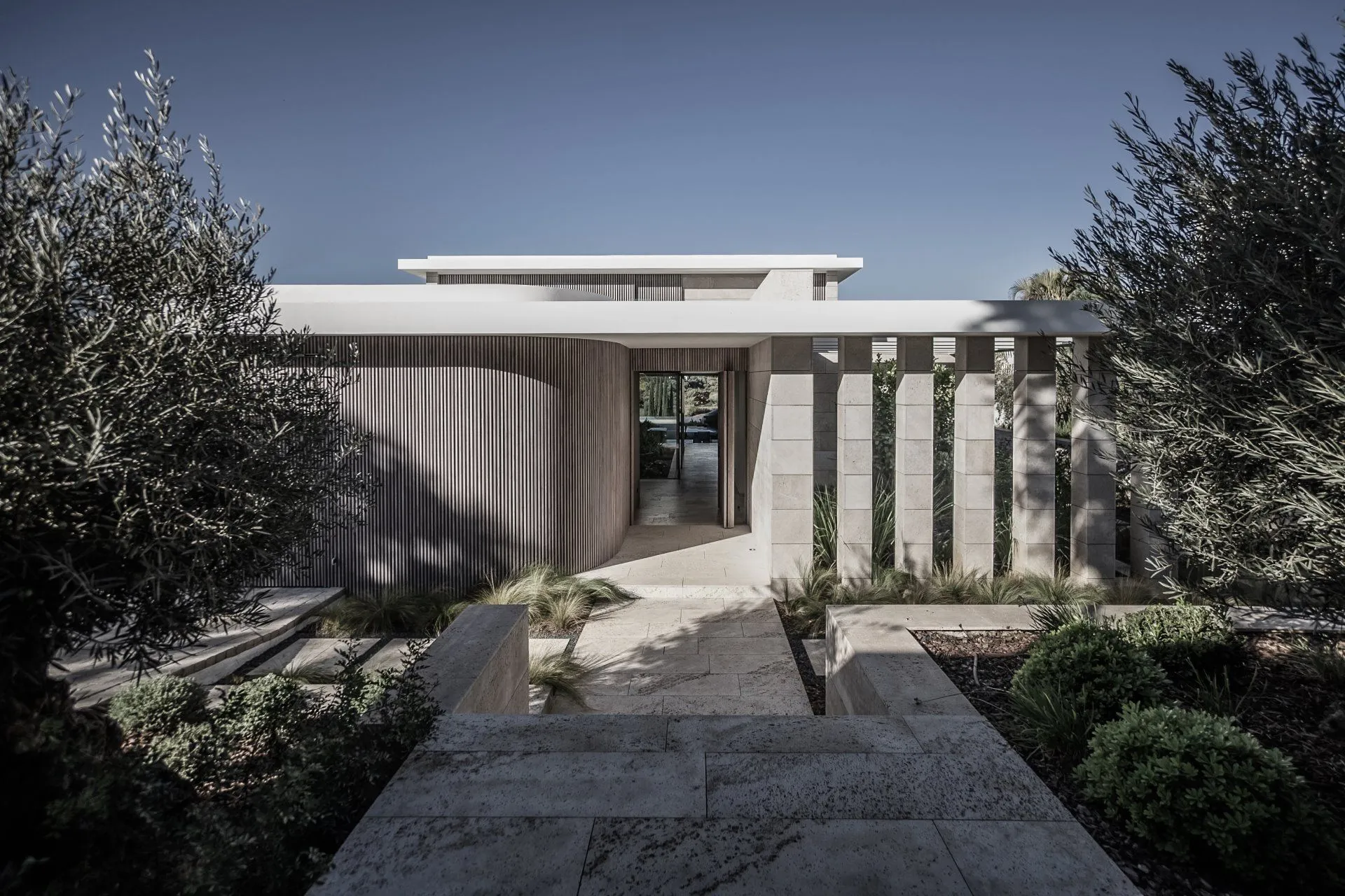 Modern villa entrance featuring travertine stone columns, curved wood cladding, and Mediterranean olive trees.