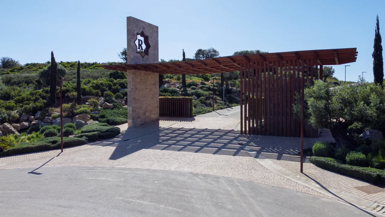 Modern entrance gate with a stone pillar, metal roof casting geometric shadows. Lush green landscaping surrounds the area under a clear blue sky.