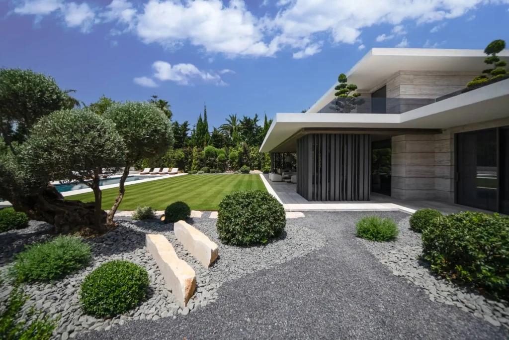 Villa Tierra exterior facade in Sotogrande by ARK Architects, featuring white stone walls, landscaped gravel garden, olive trees, and infinity pool under blue sky